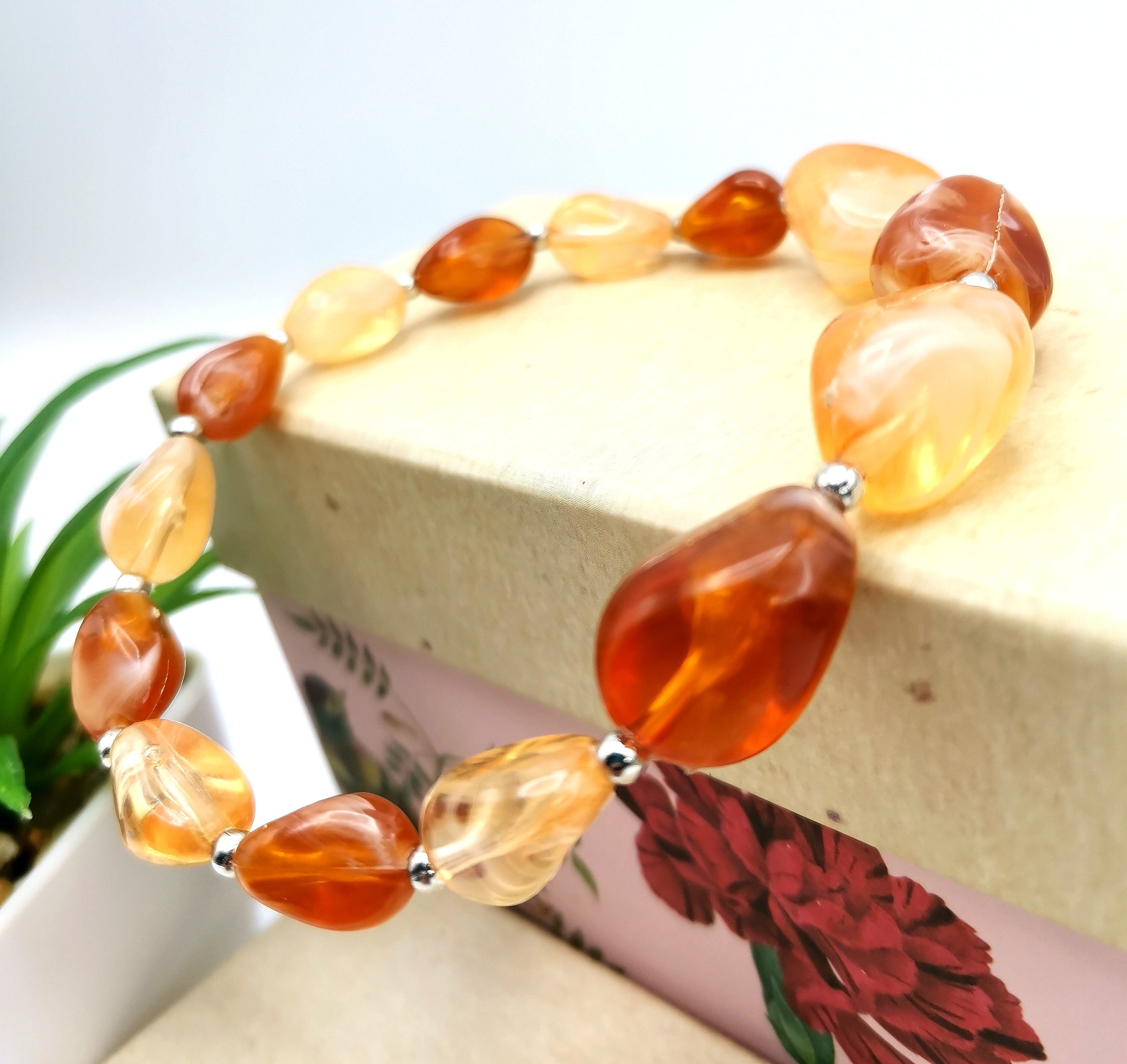 An Amber Resin Bracelet by Silver Sunbird, featuring polished amber and orange beads adorned with silver spacer beads, is elegantly displayed over a decorative box. The box, adorned with a floral pattern, sits prominently in the foreground, while a small plant provides a natural touch in the background.