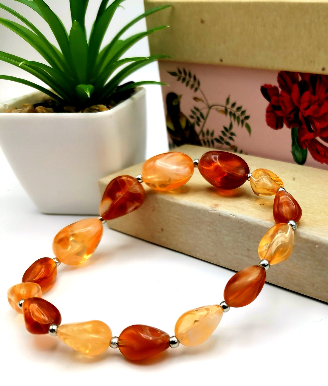 An Amber Resin Bracelet by Silver Sunbird, featuring silver spacer beads, rests on a white surface beside a small potted succulent and decorative boxes adorned with floral and bird designs.