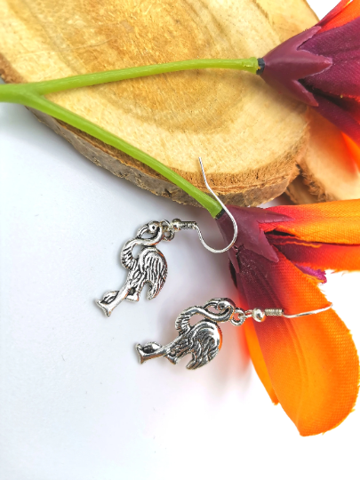 The Flocking Flamingo Earrings from Silver Sunbird are displayed on a wooden slice, accentuated by two vibrant orange and purple flower petals. The intricate flamingo design of this handmade 925 sterling silver jewellery stands out beautifully against the white background.