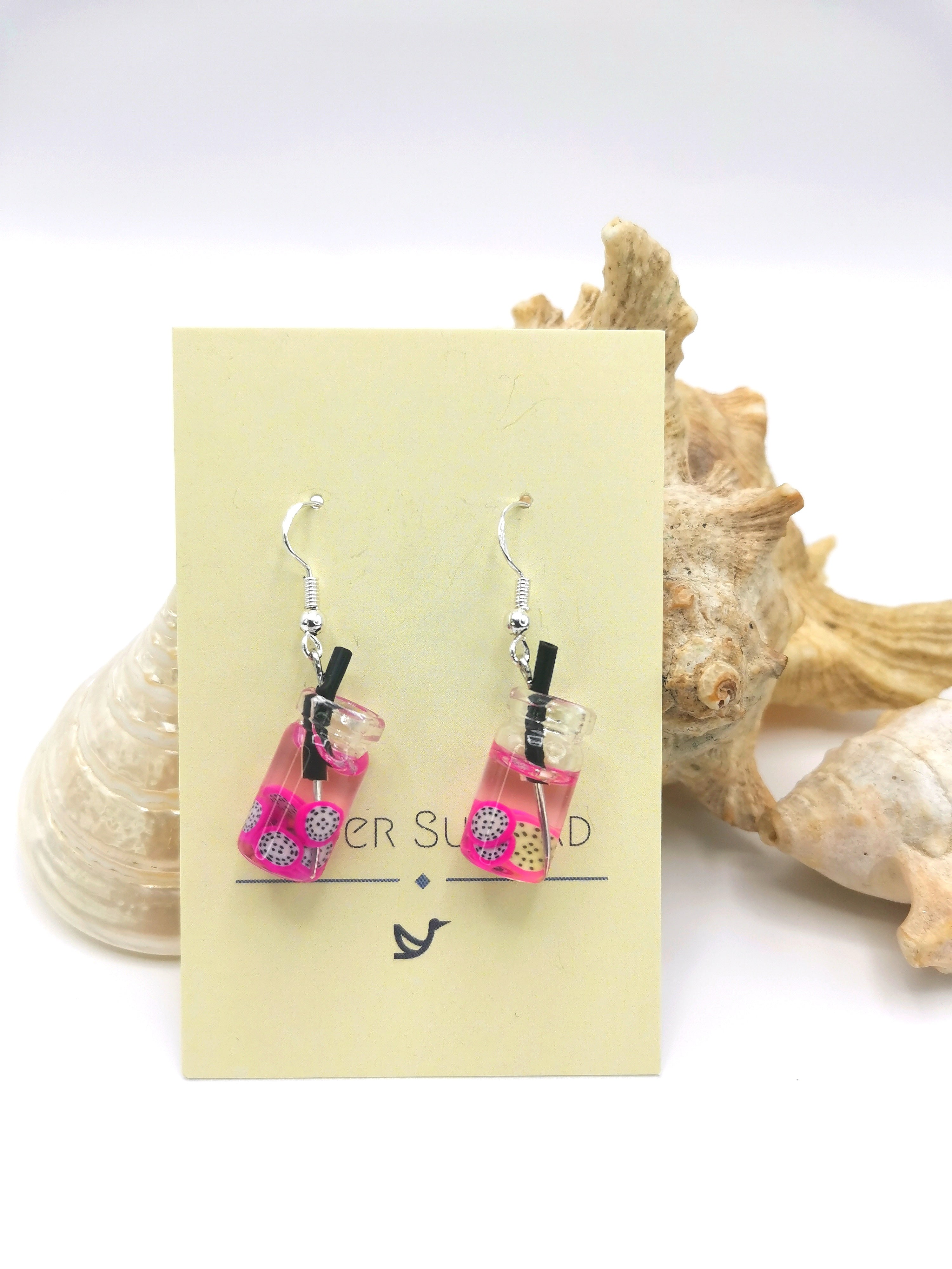 The Party Cocktail Earrings by Silver Sunbird, featuring pink and clear resin with white flowers and black strips, dangle from a "Sugar Sugar" card. Perfect for summer, they sit beautifully against seashell-adorned white surfaces.