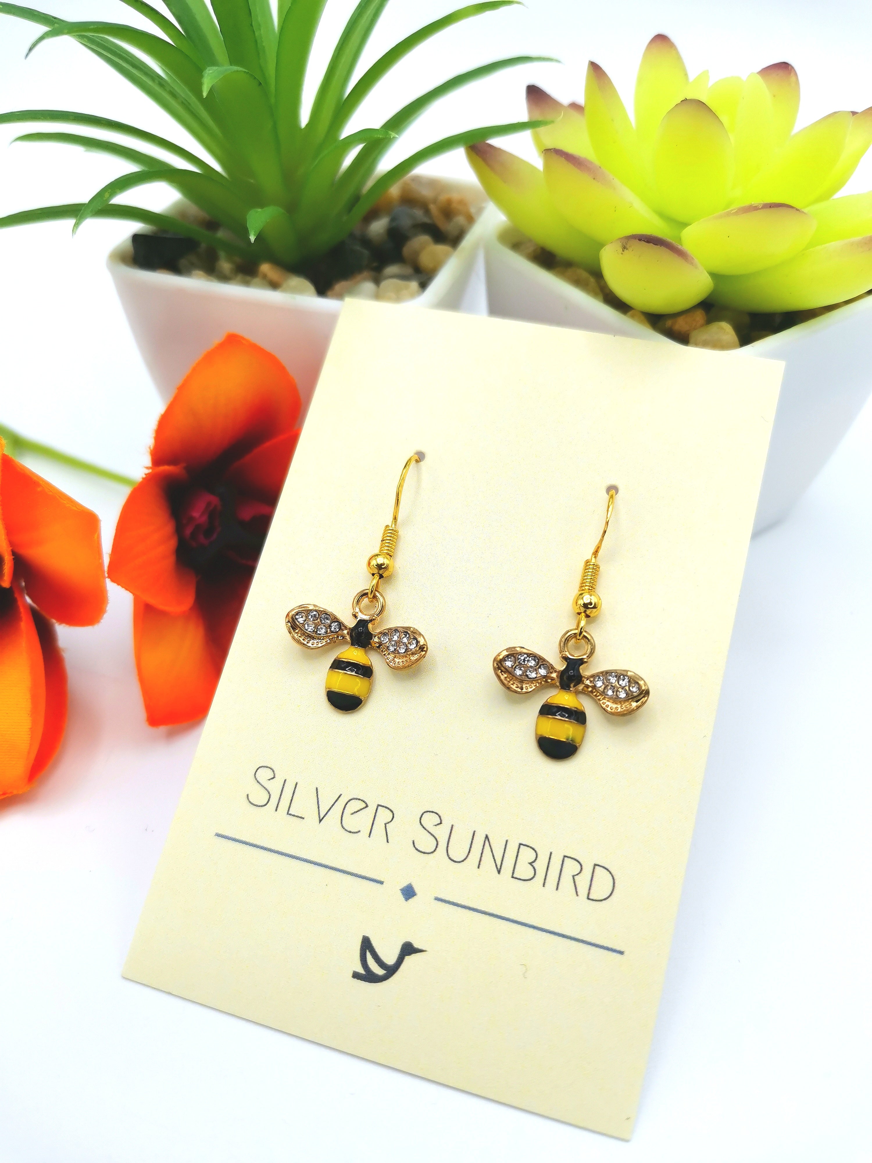 The Bee Happy Bee Earrings by Silver Sunbird are beautifully presented on a branded card. These handmade earrings showcase black and gold stripes, rhinestone wings, and sterling silver accents, all elegantly arranged among green succulents and bright orange flowers.