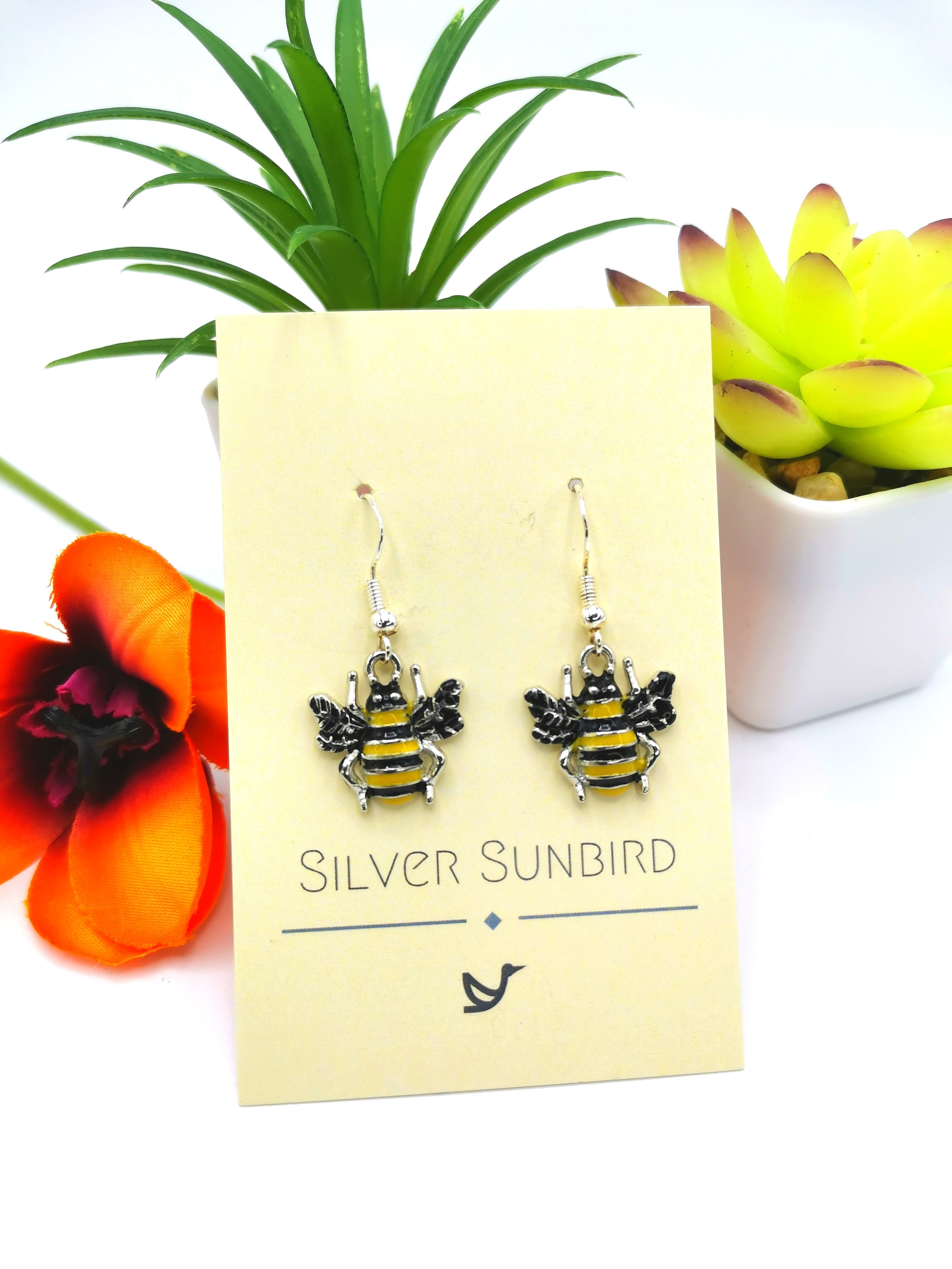 The Bee Happy Bee Earrings from Silver Sunbird are beautifully showcased on a branded card. Made from sterling silver, they are accompanied by two potted plants and an orange flower, adding a touch of color and vibrancy to the scene.