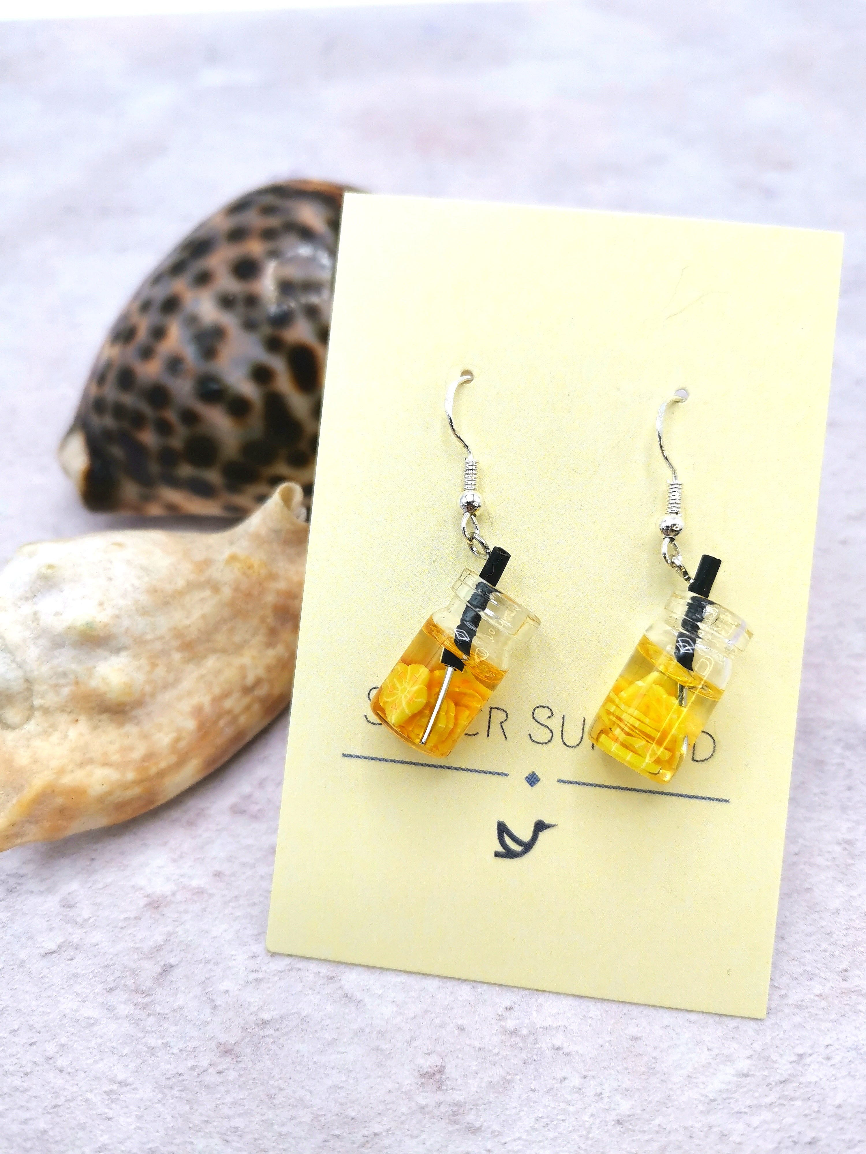 Two handmade sterling silver earrings, designed as small drinks with yellow liquid and black straws, hang on a card labeled "Silver Sunbird." The Party Cocktail Earrings rest on a light-colored surface with seashells, perfectly capturing the essence of summer.