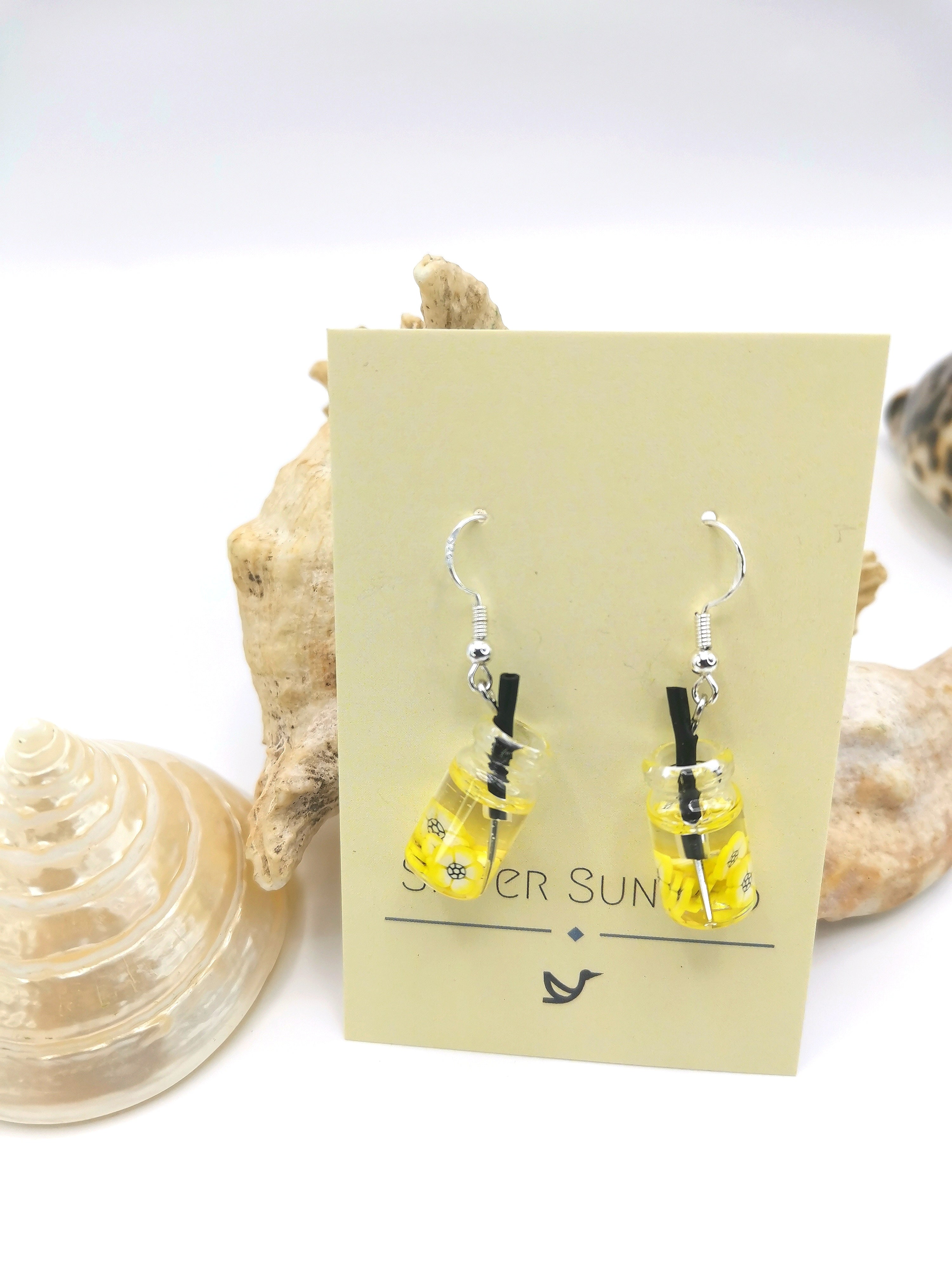 These handcrafted Party Cocktail Earrings from Silver Sunbird, resembling tiny mason jars with yellow liquid and black stir sticks, capture the essence of summer. They hang from a yellow "Silver Sun" card amidst seashells.