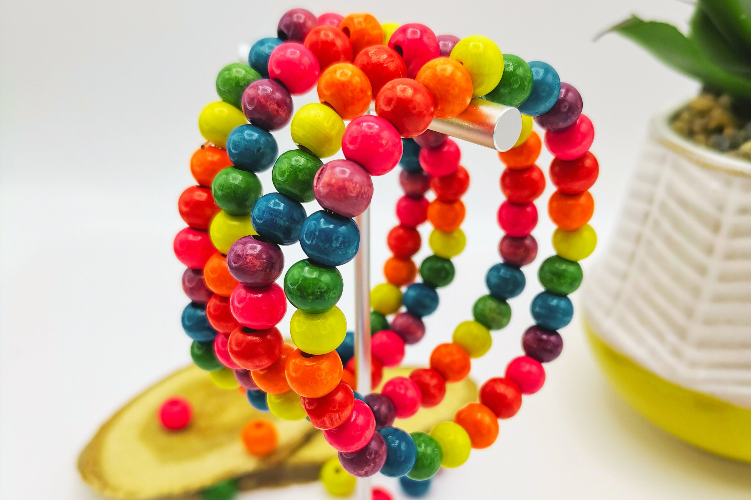 Colorful beaded bracelets with bright pink, orange, green, blue, and yellow beads displayed on a silver stand. A plant and decorative wood piece are visible in the background.