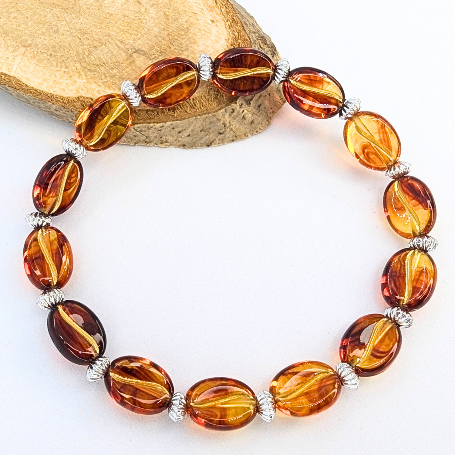 The Silver Sunbird Coffee Bean Bracelet showcases oval amber beads and silver spacers, arranged in a circle on a white surface with wood in the background. Ideal for coffee enthusiasts wanting to mix their love for coffee with stylish accessories.