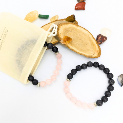 Two Rose Quartz and Lava Stone Bracelets from Silver Sunbird are displayed on a surface with a beige drawstring pouch partially visible. Surrounding them are various polished stones and wooden slices.