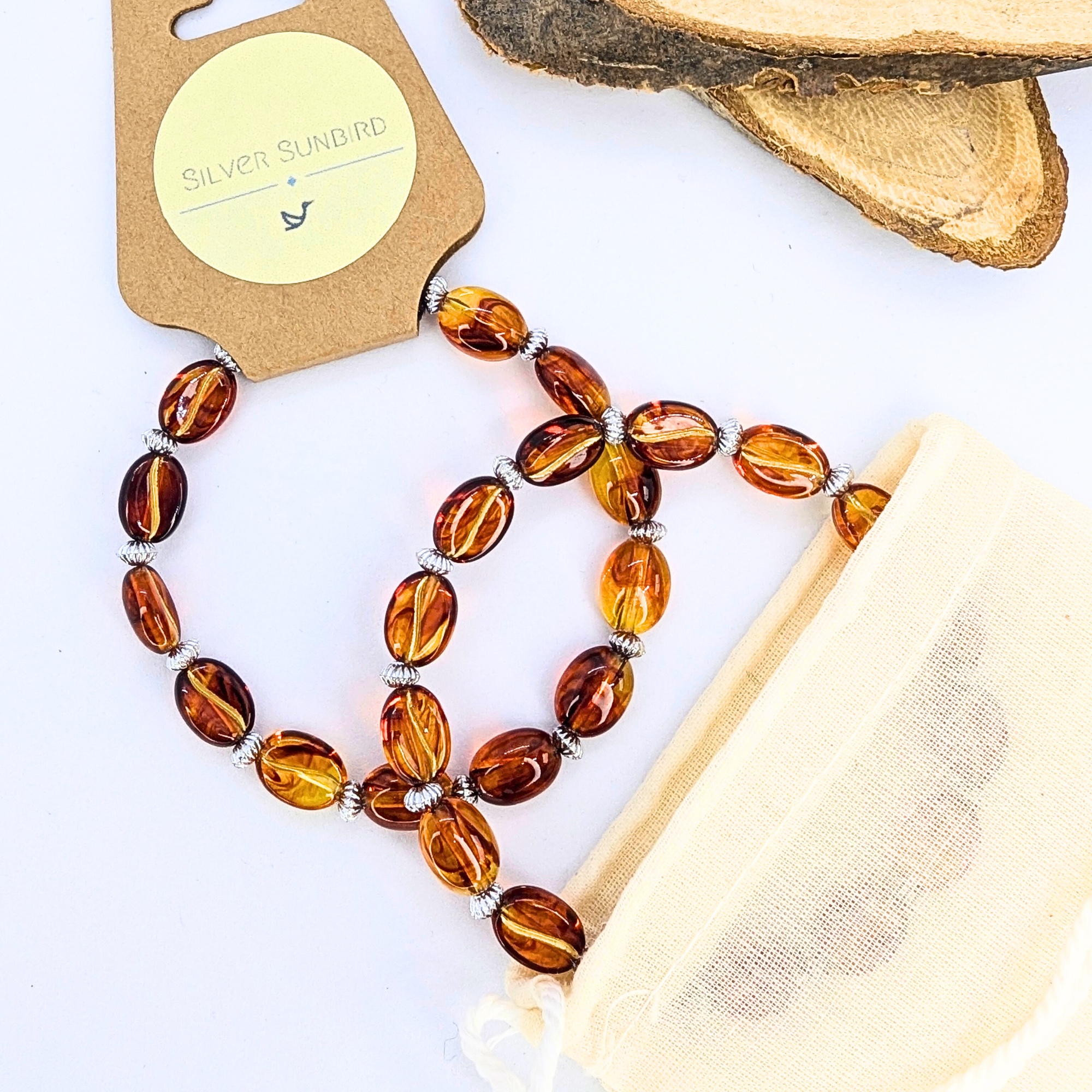 Displayed on a white surface, an amber beaded necklace with silver spacers lies beside a beige &quot;Silver Sunbird&quot; pouch. Wood slices in the background add charm reminiscent of Silver Sunbird&