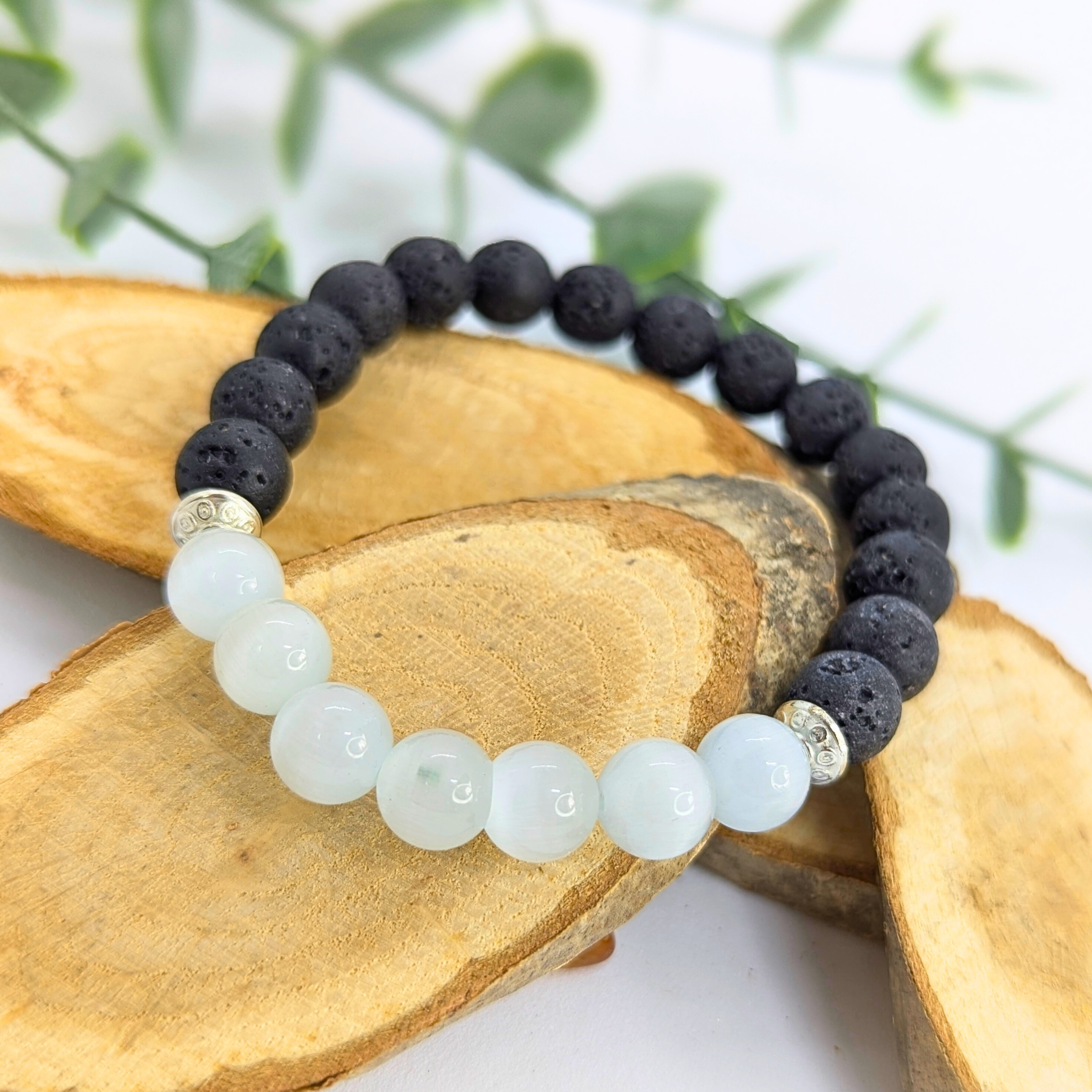 The Silver Sunbird Moonstone and Lava Bracelet, featuring black lava stones, white moonstone beads, and metallic spacers, is artfully displayed on wooden slices with green leaves in the background.