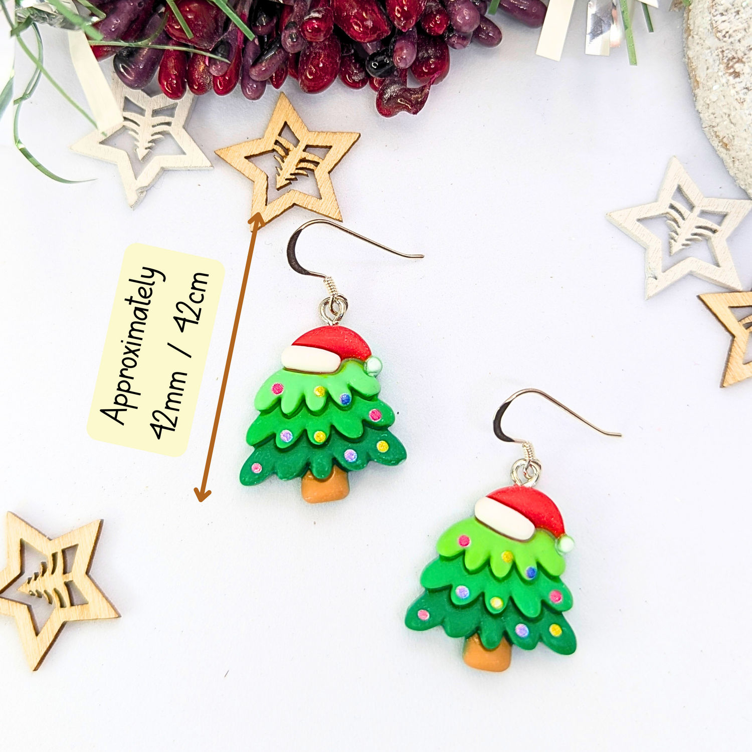 Adorned with vibrant red Santa hats and colorful decorations, the Christmas Tree Earrings by Silver Sunbird make for the perfect holiday accessory. Displayed on a white surface and surrounded by star-shaped wooden decorations, these festive jewelry pieces measure approximately 42mm / 12cm.
