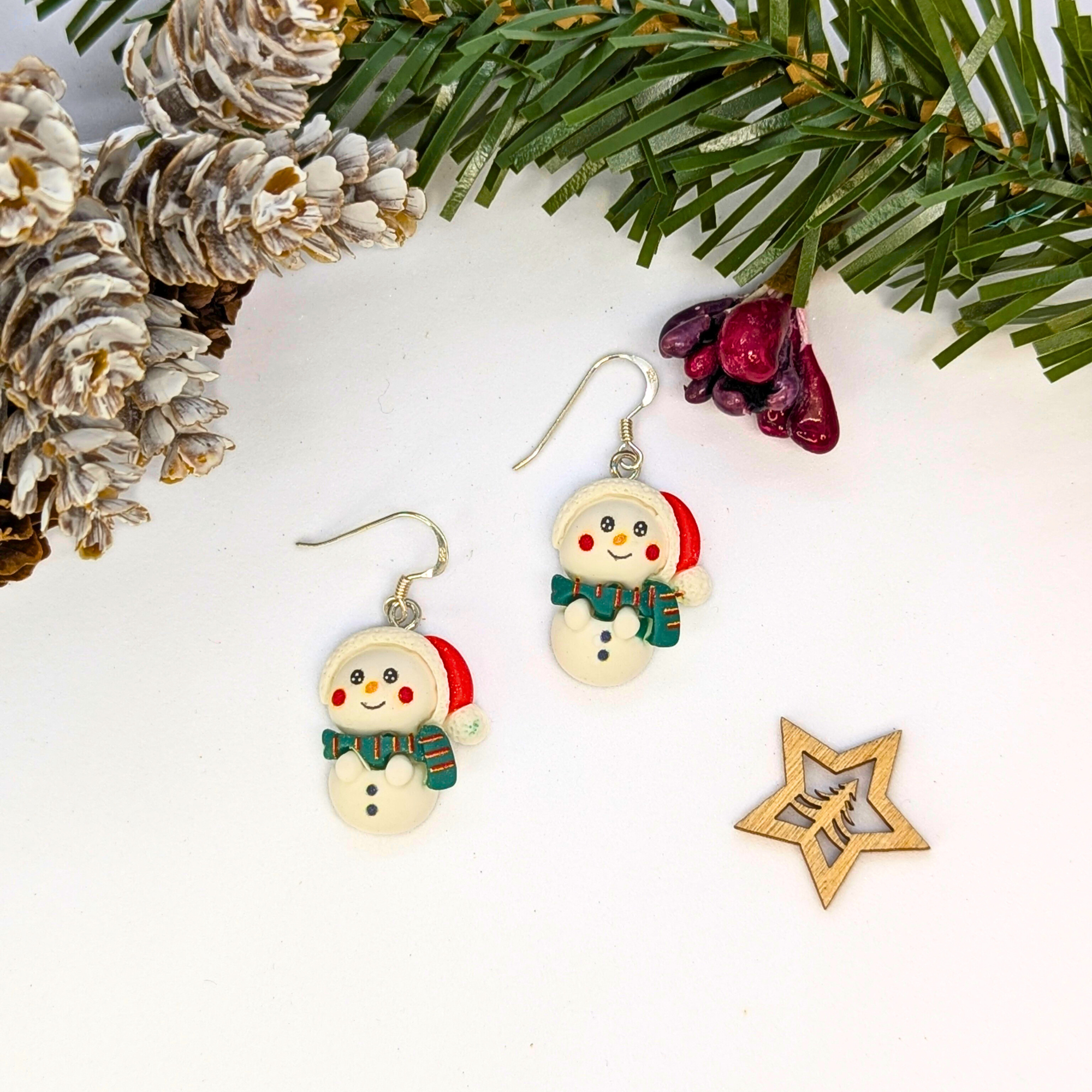 The Bright Snowmen Earrings by Silver Sunbird, featuring red hats and green scarves, are shown on a white surface with pine decor—perfect festive accessories for your Christmas jewelry collection.