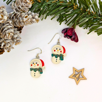 The Bright Snowmen Earrings by Silver Sunbird, featuring red hats and green scarves, are shown on a white surface with pine decor—perfect festive accessories for your Christmas jewelry collection.