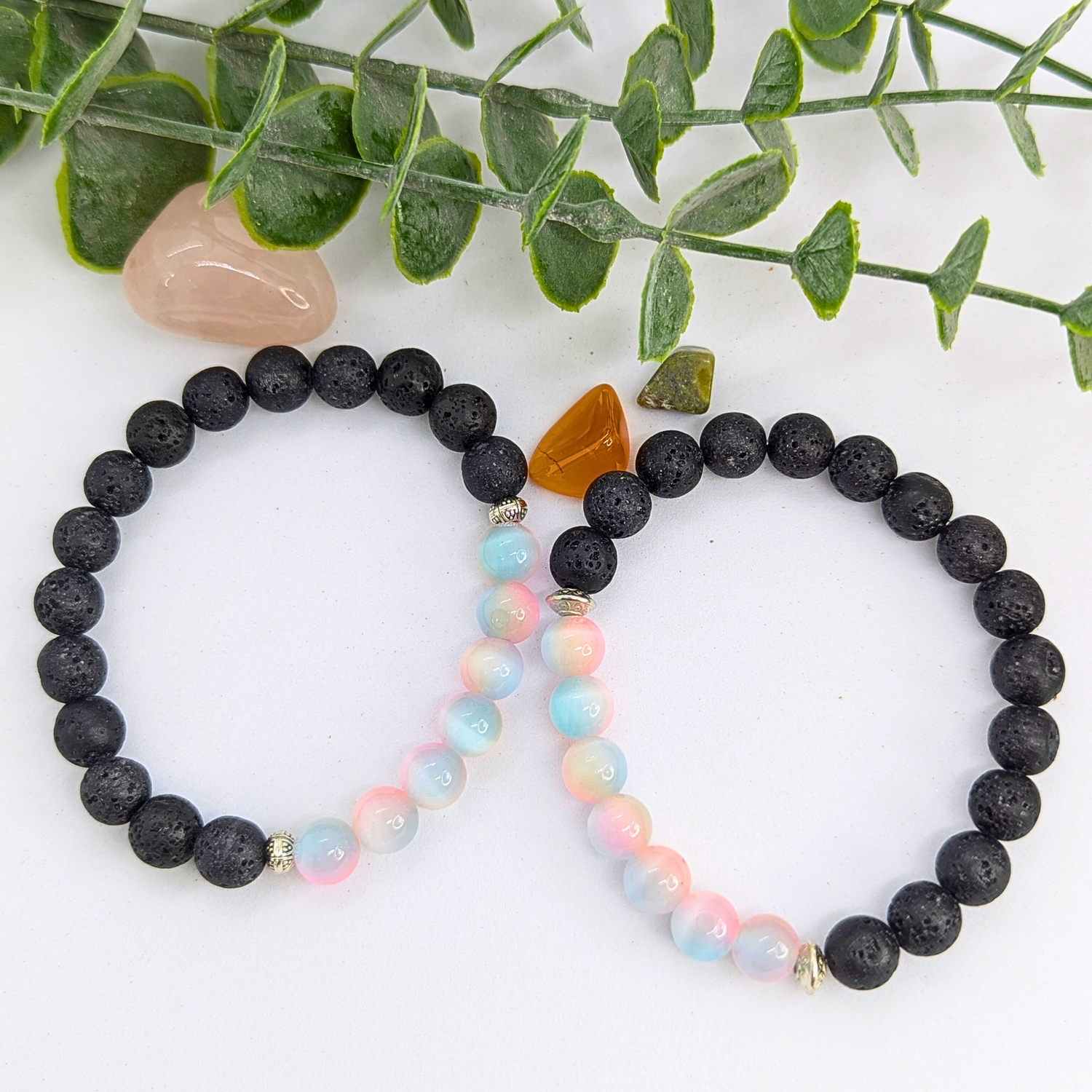 A pastel beaded bracelet and the Silver Sunbird Mermaid-Inspired Lava Stone Bracelet with black beads and a brownish-orange stone rest on a white surface, with green leaves and polished stones in the background.