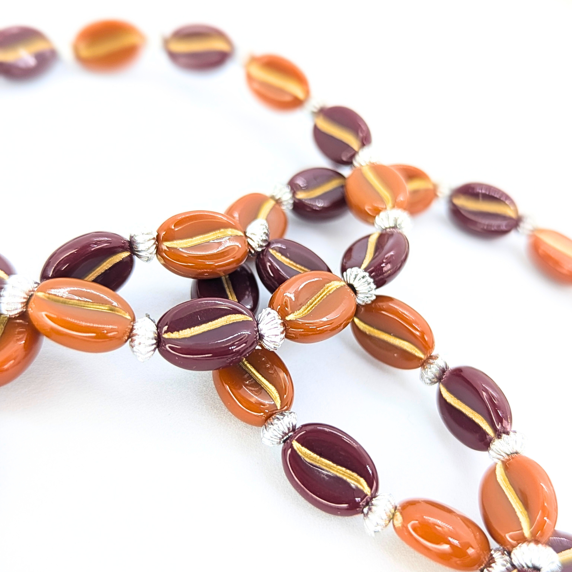 The Silver Sunbird Coffee Bean Bracelet, showcasing alternating oval beads in orange and dark red with gold stripes, separated by small silver beads, is displayed on a white surface, reflecting the rich tones cherished by coffee aficionados.