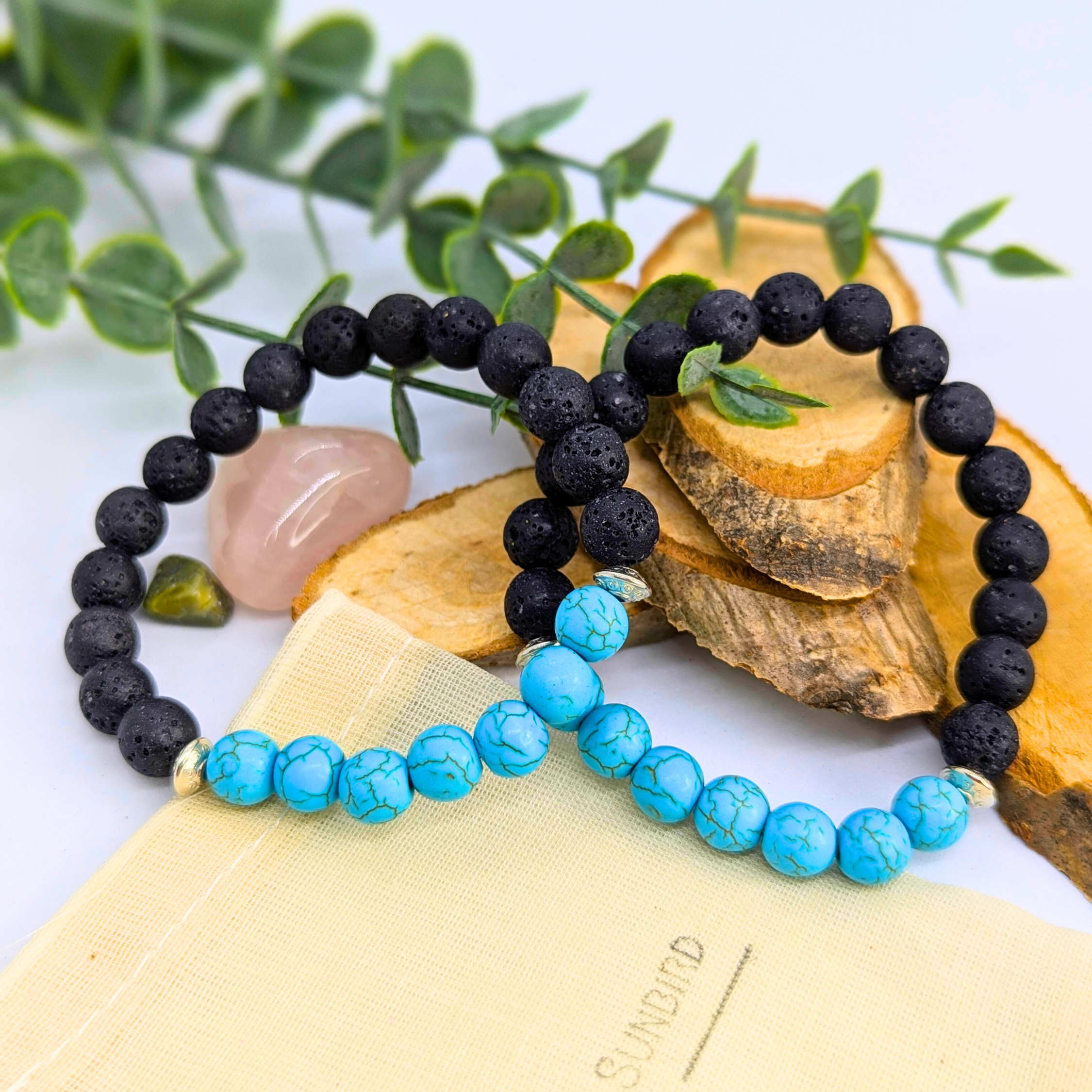 The Silver Sunbird Turquoise and Lava Stone Bracelets are displayed on wooden slices with green leaves and crystals, featuring one black lava stone bracelet and one turquoise bead bracelet, both set on a cream-colored pouch.