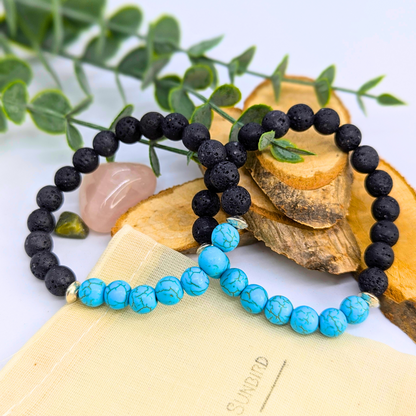 The Silver Sunbird Turquoise and Lava Stone Bracelets are displayed on wooden slices with green leaves and crystals, featuring one black lava stone bracelet and one turquoise bead bracelet, both set on a cream-colored pouch.
