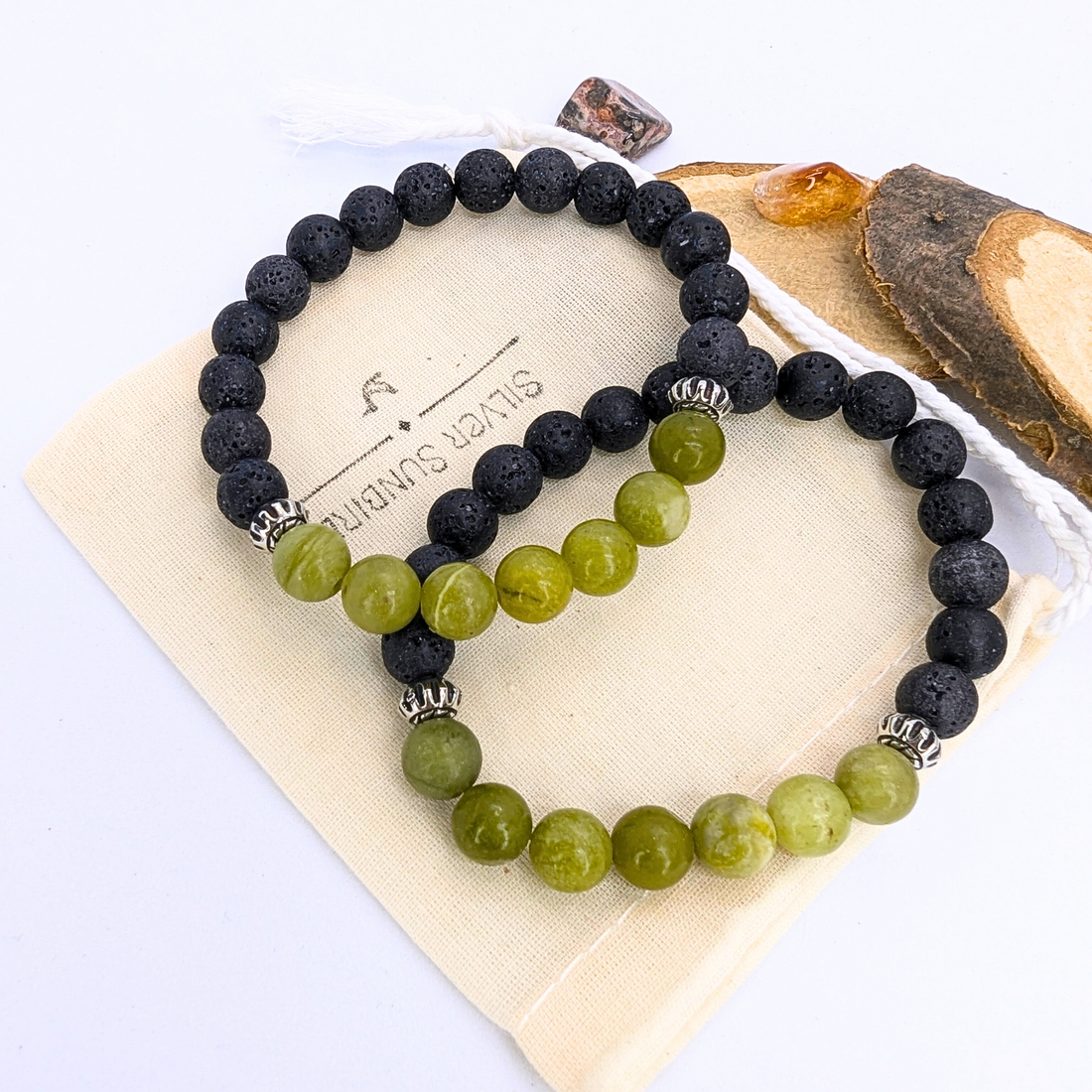 Two beaded gemstone bracelets—the Peridot and Lava Stone Bracelets by Silver Sunbird—rest on a beige fabric pouch, beside a piece of wood and two amber stones, all set against a white background.