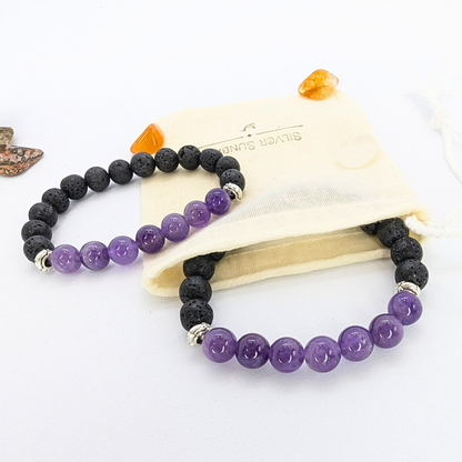 Two stretch bracelets featuring black lava stones and purple amethyst beads are displayed on a wooden slice. A beige drawstring pouch with &quot;Silver Surfer&quot; branding is positioned to the side, along with small polished stones in orange and brown hues. The amethyst beads are glossy and smooth, contrasting with the porous texture of the lava stones.