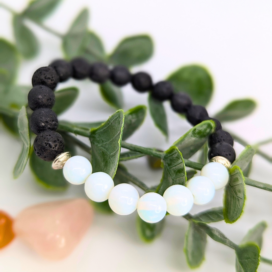 The Silver Sunbird Opal and Lava Stone Bracelet, featuring matte black lava and glossy white opal beads, is displayed on green leafy stems with a blurred pink stone in the background.