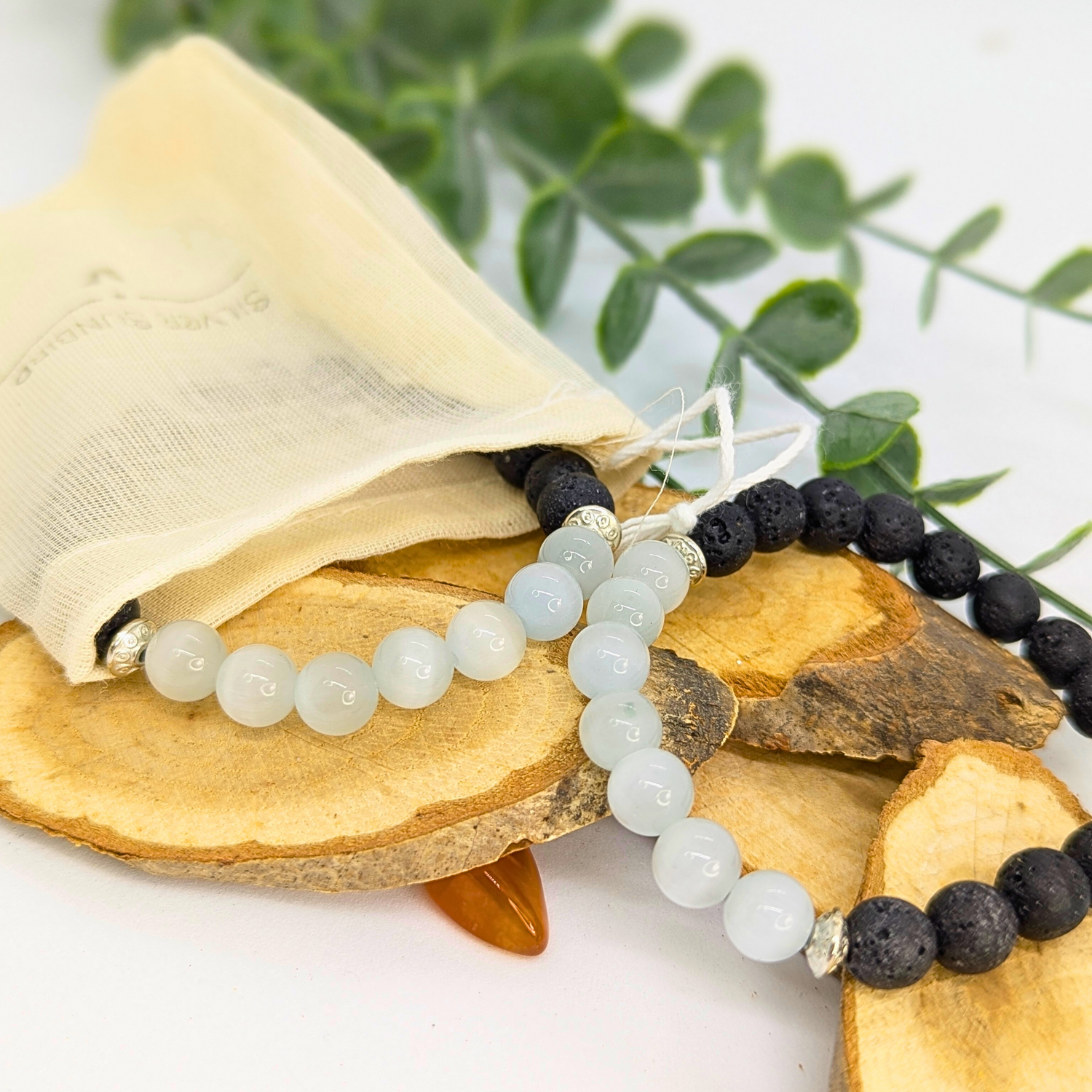 The Moonstone and Lava Bracelets from Silver Sunbird, featuring black lava stones and light blue moonstone beads, are displayed on wooden slices with a cream drawstring pouch and green leafy stem for a tranquil touch.