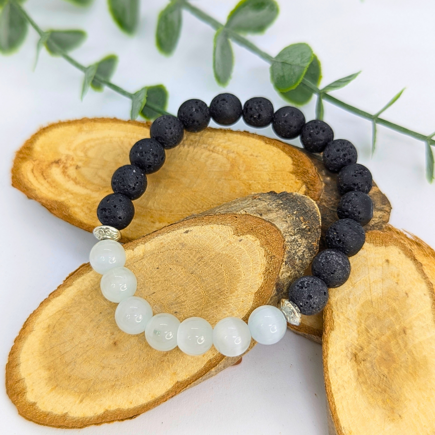 The Silver Sunbird Moonstone and Lava Bracelets are showcased on three overlapping wooden slices, accented with green leaves in the background, all placed on a white surface.