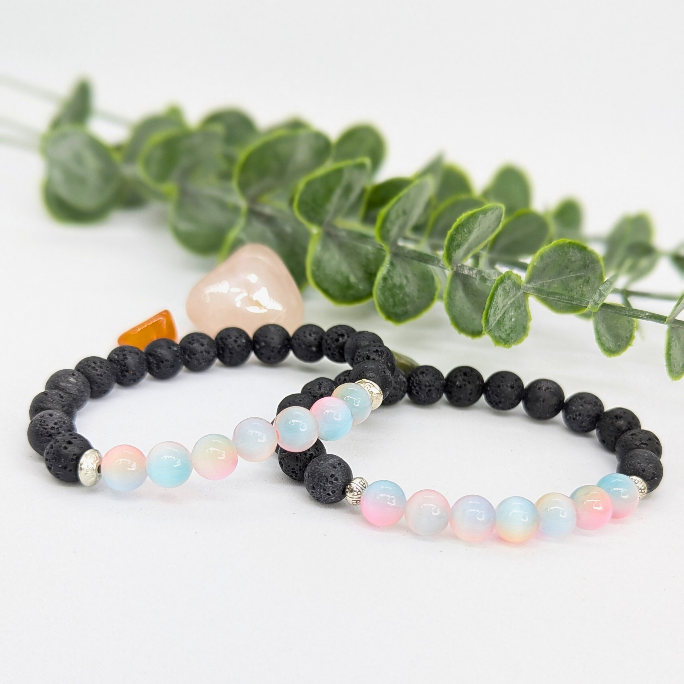 Two beaded bracelets sit before green eucalyptus: the Silver Sunbird Mermaid-Inspired Lava Stone Bracelet with black beads and a pink stone, and a pastel bracelet with black accents—ideal gifts for ocean lovers.