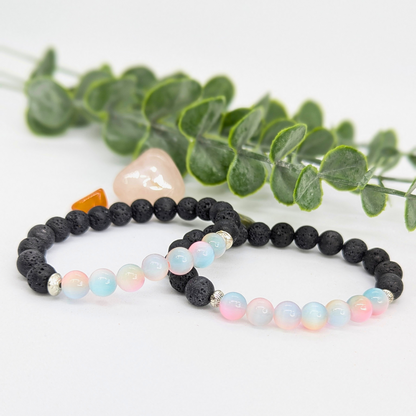 Two beaded bracelets sit before green eucalyptus: the Silver Sunbird Mermaid-Inspired Lava Stone Bracelet with black beads and a pink stone, and a pastel bracelet with black accents—ideal gifts for ocean lovers.