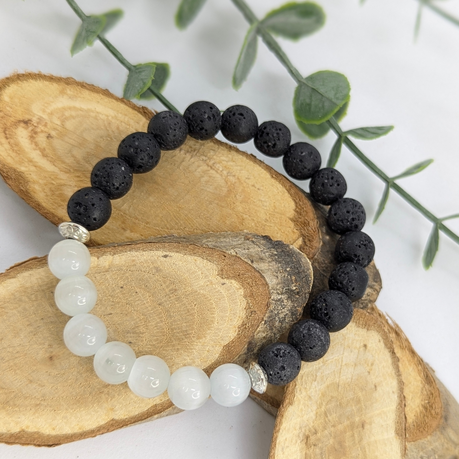 The Silver Sunbird Moonstone and Lava Bracelet, handcrafted with half black lava stones, half white moonstone beads, and two silver spacers, is displayed on oval wooden slices with green leaves in the background.