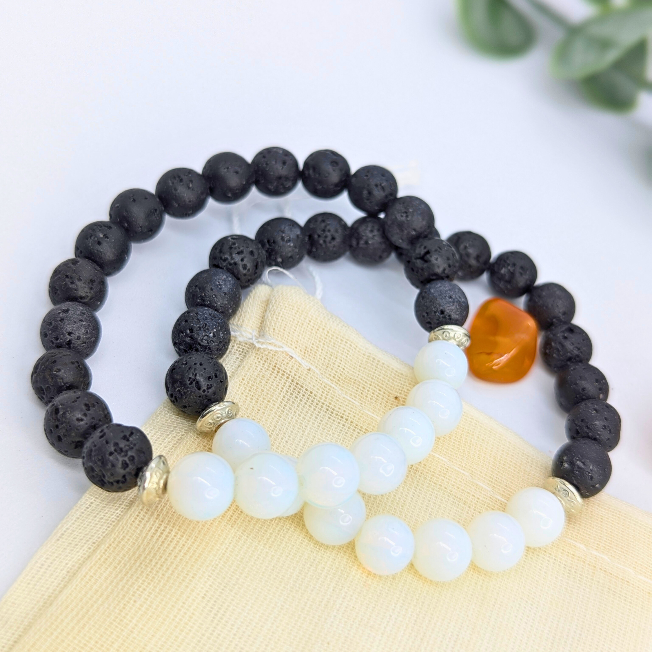Two Silver Sunbird Opal and Lava Stone Bracelets rest on a beige pouch, one with black lava beads and the other with white opal beads, both accented by silver spacers. An orange stone and green leaves appear in the background.