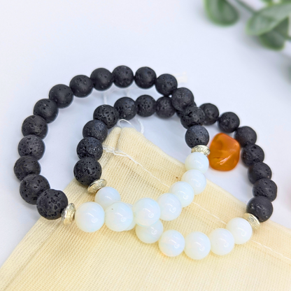 Two Silver Sunbird Opal and Lava Stone Bracelets rest on a beige pouch, one with black lava beads and the other with white opal beads, both accented by silver spacers. An orange stone and green leaves appear in the background.