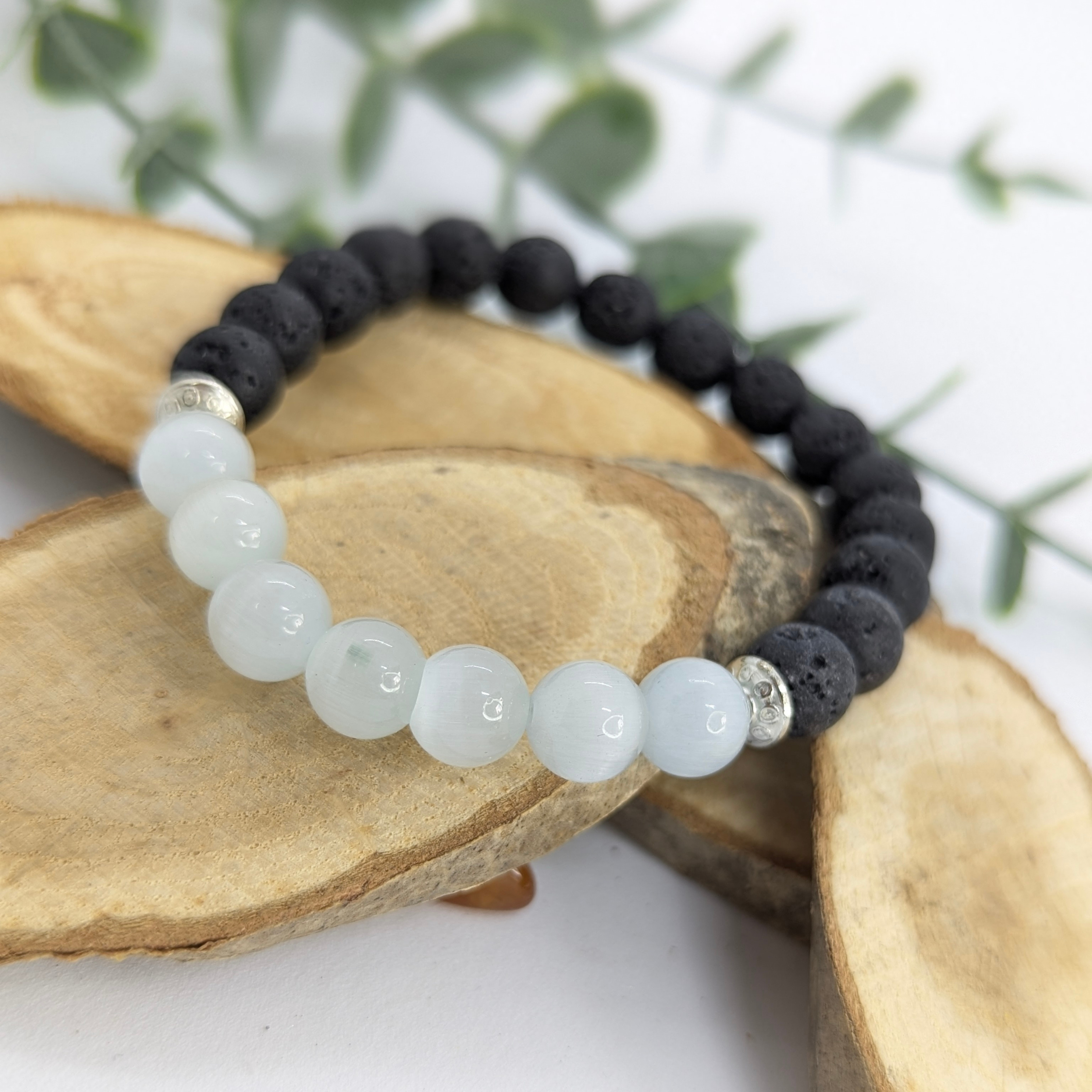 The Silver Sunbird Moonstone and Lava Bracelets, featuring black lava stone and light blue moonstone beads, are displayed on stacked wooden slices with a blurred green leafy background.