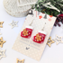 Gingerbread Men Stocking Earrings by Silver Sunbird are showcased on a cardboard backing, accompanied by wooden star ornaments and a snowy decorative branch.