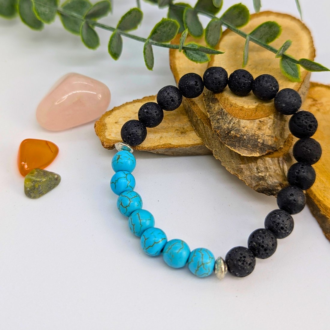 The Silver Sunbird Turquoise and Lava Stone Bracelets are shown on wooden slices, accompanied by a sprig of green leaves and three polished stones in peach, orange, and green.