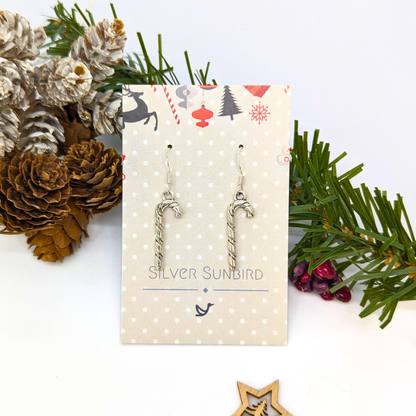 Classic Candy Cane Earrings by Silver Sunbird hang from a branded card, surrounded by pinecones, evergreen branches, and festive holiday décor—perfect Christmas jewelry for the season.