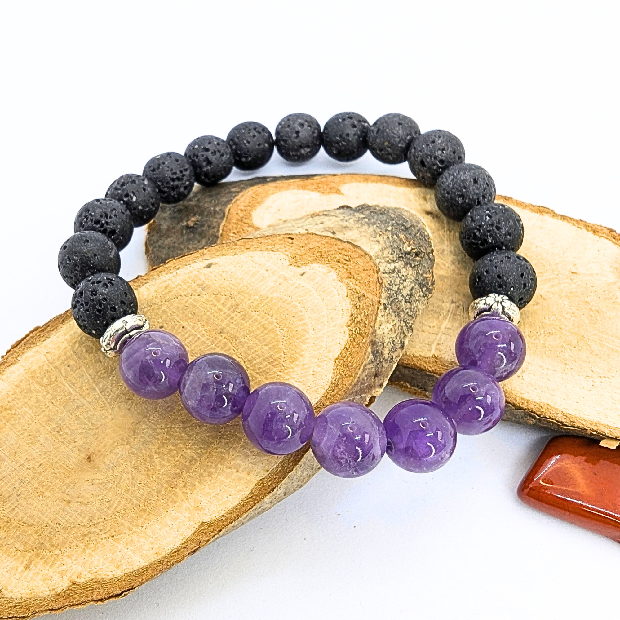 A stretch bracelet made with black lava stones and polished purple amethyst beads is displayed on a wooden slice. The amethyst beads are glossy and vibrant, contrasting with the textured, porous appearance of the lava stones. A small orange polished stone is visible in the background, adding a touch of colour to the composition.