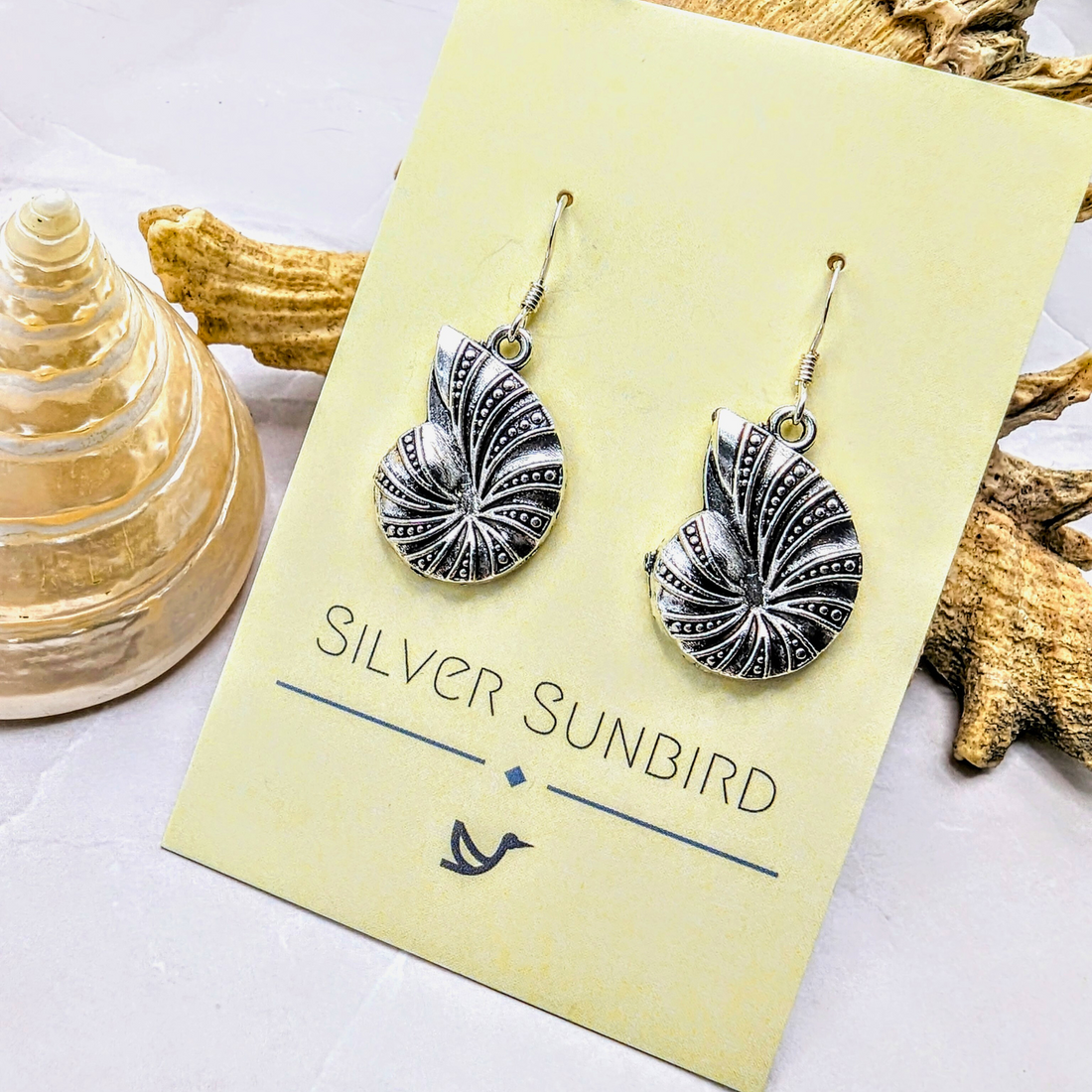 A pair of Shoreline Spiral Shell Earrings by Silver Sunbird, displayed on a branded card beside a spiral seashell and driftwood, evokes the elegance of ocean-inspired jewelry on a light backdrop.
