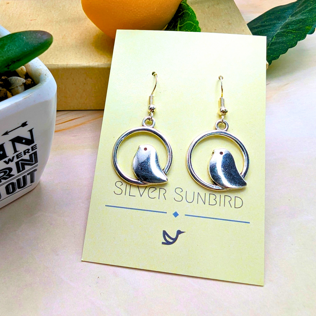 Displayed on a Silver Sunbird card, the Beautiful Bird Earrings are crafted from 925 Sterling Silver, showcasing a delicate bird in circular frames. A small plant and decor enhance the elegant presentation, perfect for bird lovers.