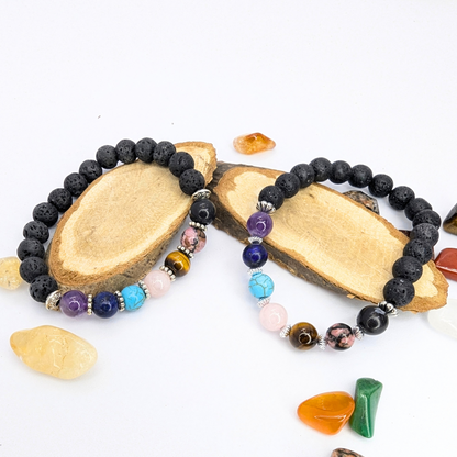 Two Silver Sunbird The Chakra Bracelets with black lava stones and colorful semi-precious gemstones rest on wooden slabs, surrounded by polished multicolor stones, all set against a white background.