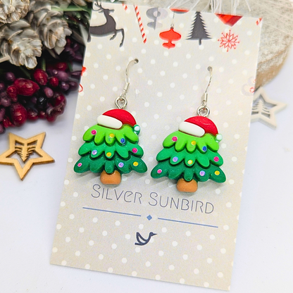 Displayed on a decorative card bearing the name &quot;Silver Sunbird,&quot; these festive Christmas Tree Earrings are embellished with a Santa hat and colorful ornaments. This charming holiday accessory is presented amidst seasonal decorations and an elegant pinecone arrangement.