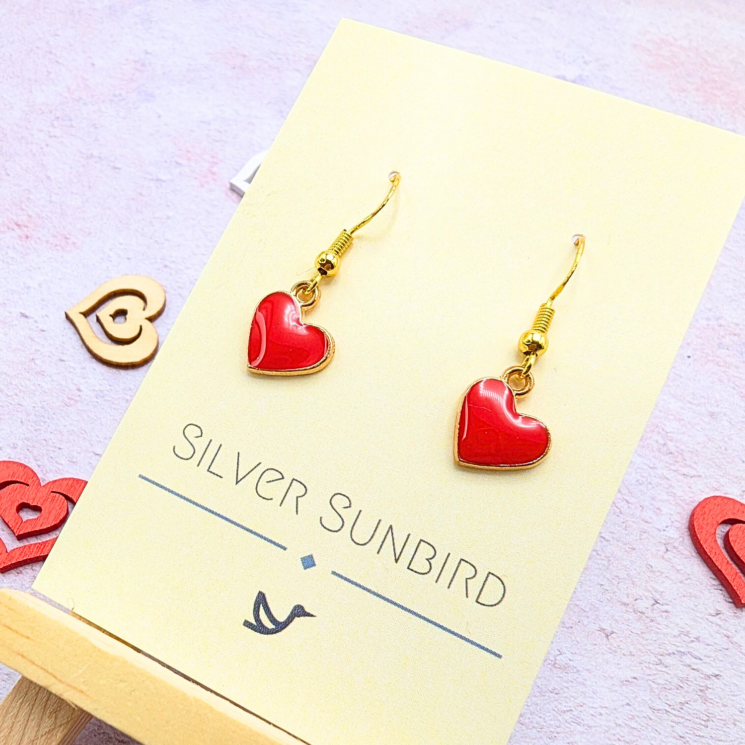 The Alluring Red Heart Earrings, by Silver Sunbird, are golden with red heart-shaped pendants displayed on a yellow card. A pink background with heart motifs enhances the charm of these hypoallergenic earrings.