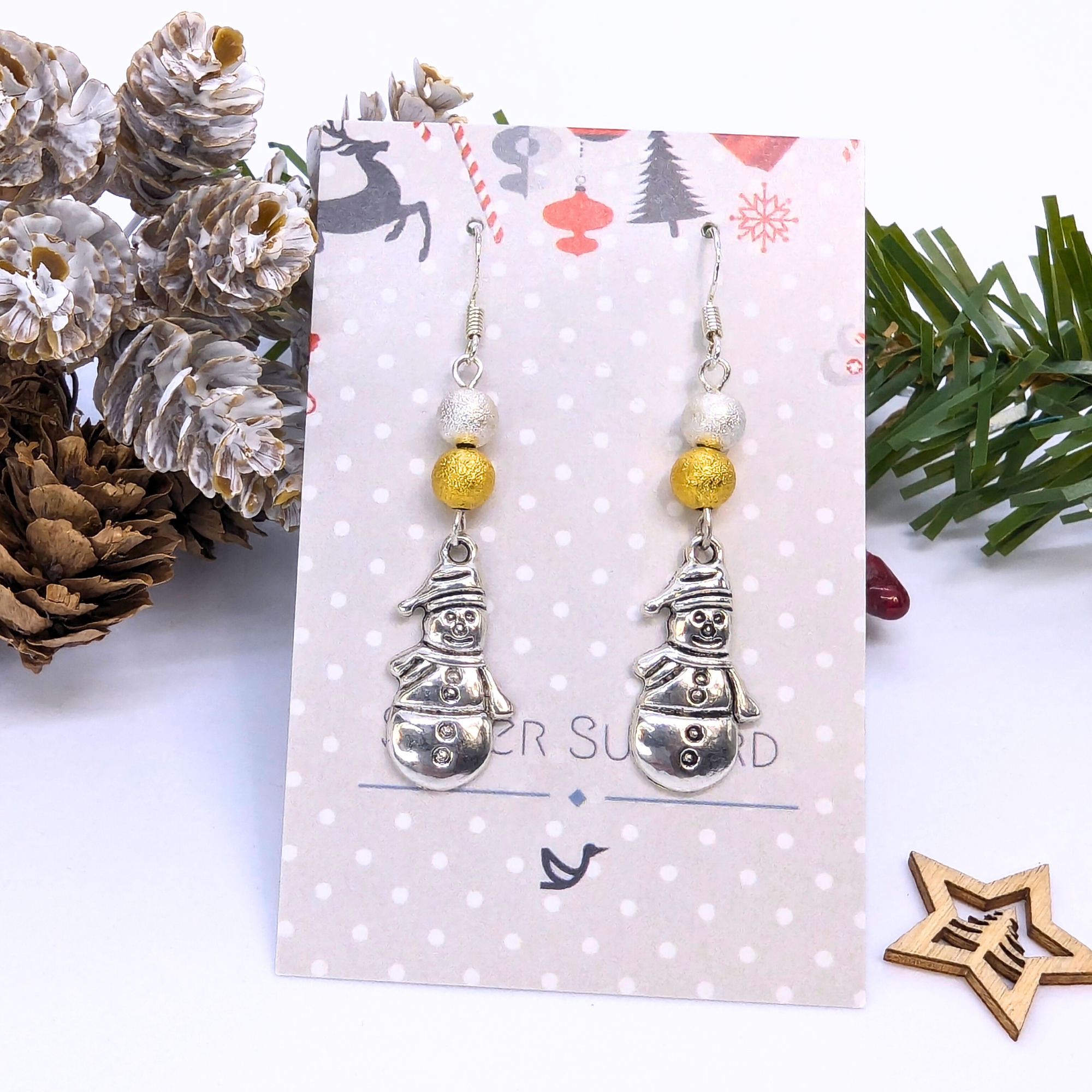 The Very Merry Snowmen Earrings by Silver Sunbird, crafted with gold beads and sterling silver, are displayed on a festive card surrounded by pine cones, greenery, and a wooden star for a charming holiday presentation.