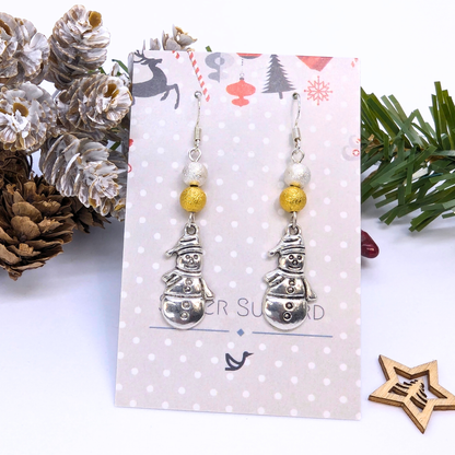 The Very Merry Snowmen Earrings by Silver Sunbird, crafted with gold beads and sterling silver, are displayed on a festive card surrounded by pine cones, greenery, and a wooden star for a charming holiday presentation.