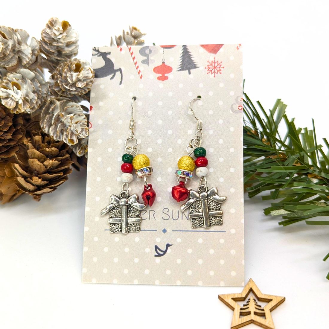 Gift Box Dangle Earrings by Silver Sunbird feature silver present charms, colorful beads, and red heart accents on sterling silver hooks, displayed on a polka dot card with pinecones, evergreen branches, and a wooden star.
