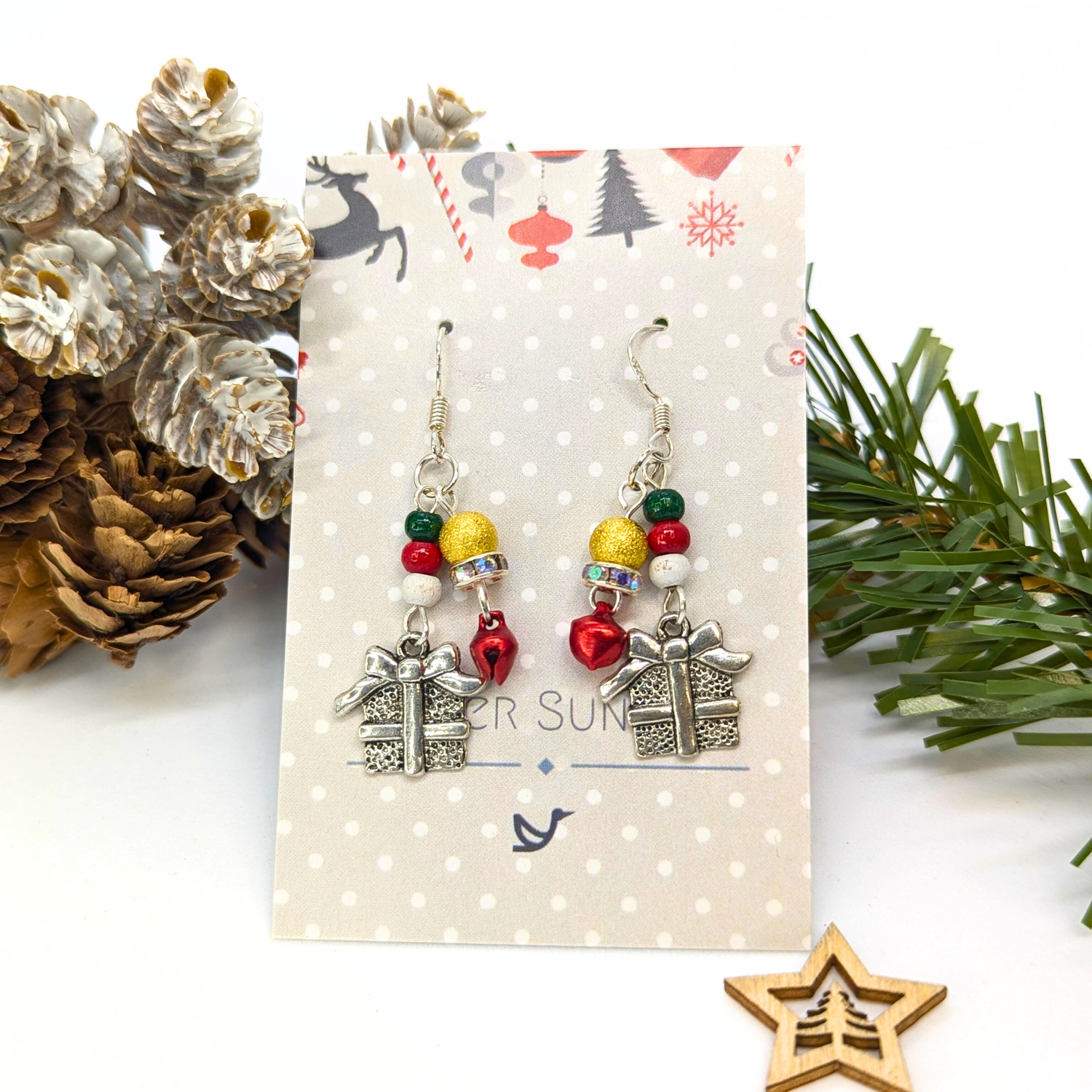 Gift Box Dangle Earrings by Silver Sunbird feature silver present charms, colorful beads, and red heart accents on sterling silver hooks, displayed on a polka dot card with pinecones, evergreen branches, and a wooden star.