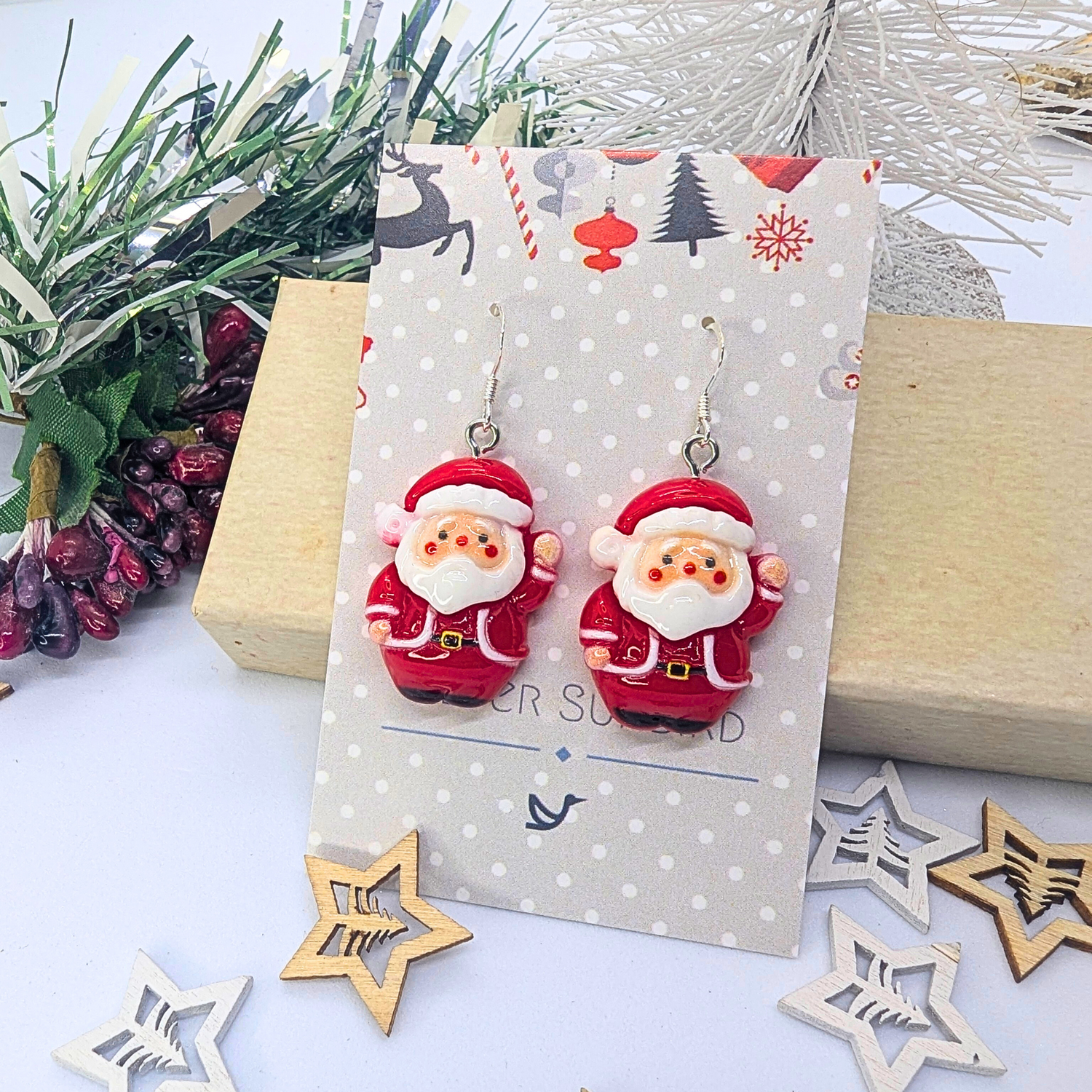 The Jolly Santa Earrings from Silver Sunbird are showcased on a decorative card, surrounded by holiday-themed decor featuring a small tree, pine branches, and wooden stars. This setup creates a cheerful Christmas atmosphere for these delightful festive accessories.