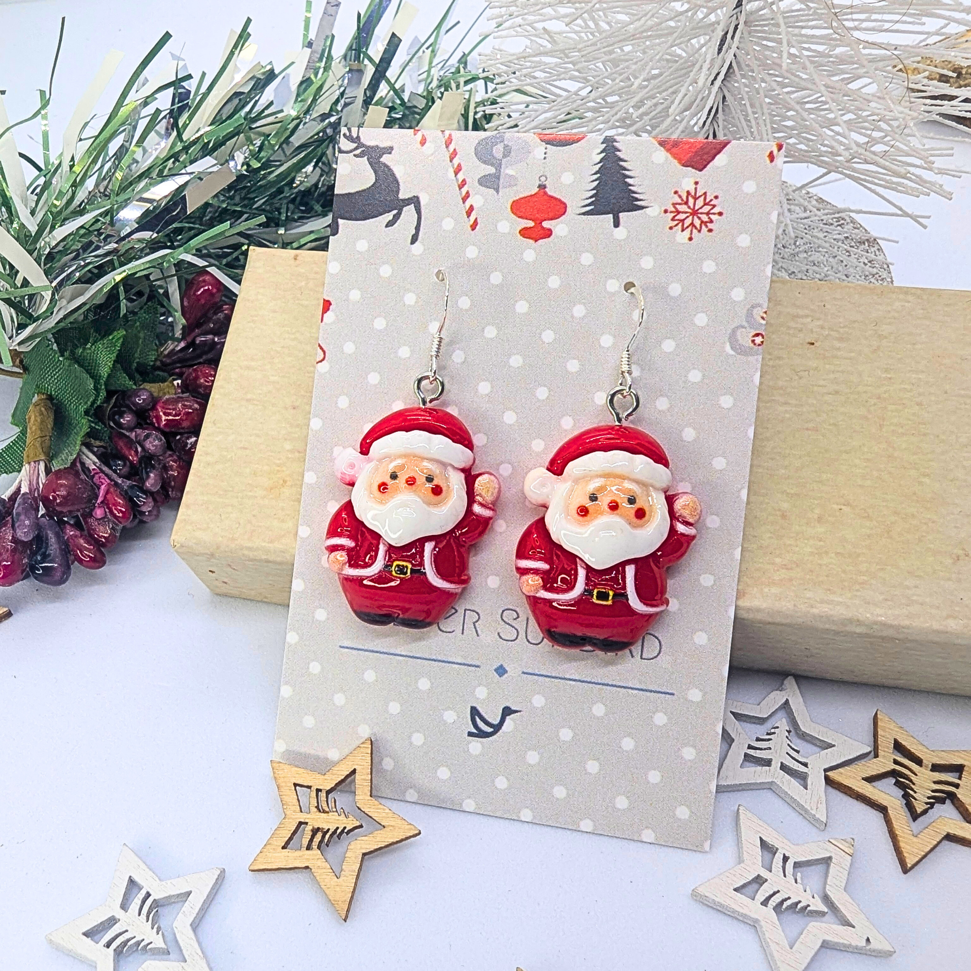 The Jolly Santa Earrings from Silver Sunbird are showcased on a decorative card, surrounded by holiday-themed decor featuring a small tree, pine branches, and wooden stars. This setup creates a cheerful Christmas atmosphere for these delightful festive accessories.