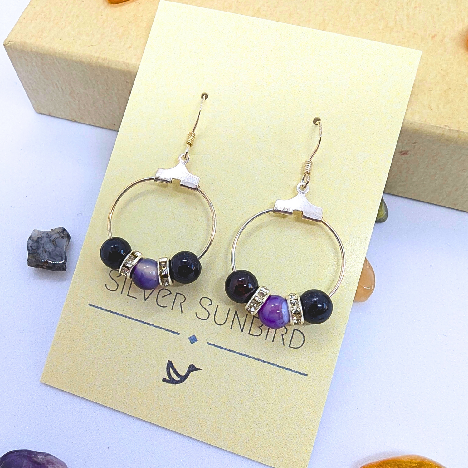 A pair of circular silver earrings embellished with purple gemstones, featuring Amethyst dangle elements and silver rhinestones, are displayed on a yellow card labelled &quot;Silver Sunbird.&quot; The card is placed on a light surface next to scattered decorative stones.