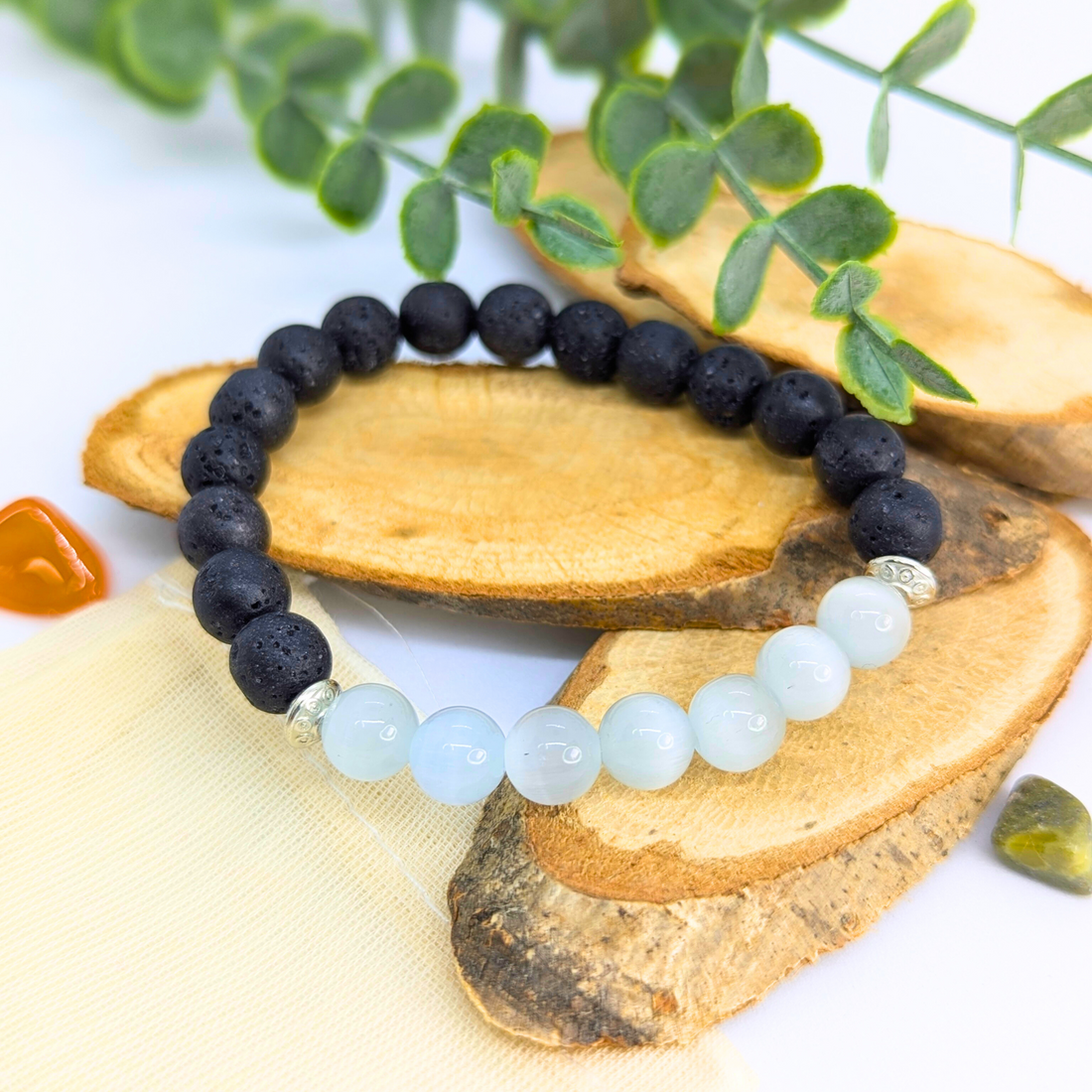 The Silver Sunbird Moonstone and Lava Bracelets are shown on wooden slices with green leaves and small polished stones, featuring handcrafted black lava beads and light blue moonstones on a white background.