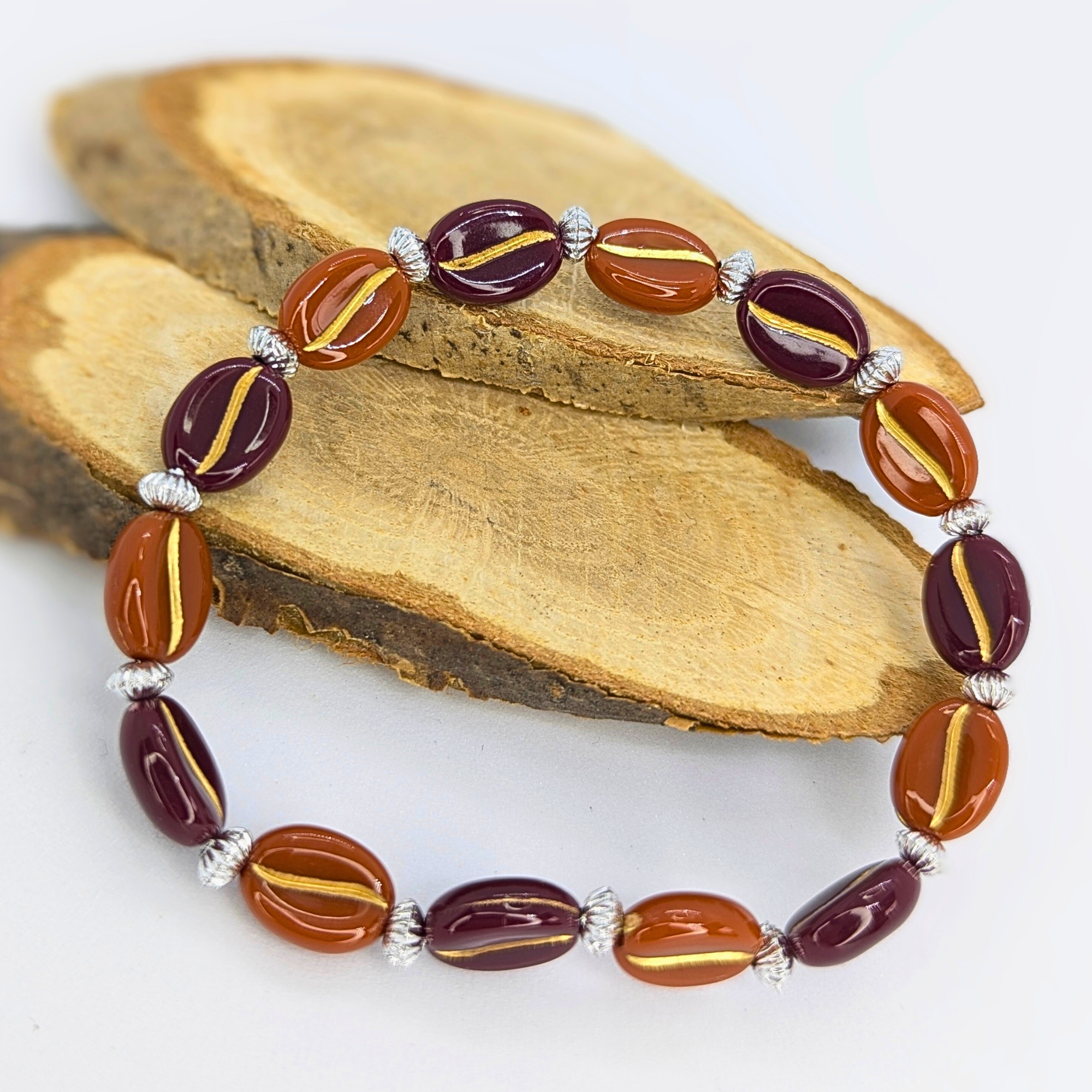 The Silver Sunbird Coffee Bean Bracelets showcase maroon and amber oval beads with gold stripes, separated by textured silver spacers. Beautifully arranged in a circle on wood, they are perfect for coffee enthusiasts.