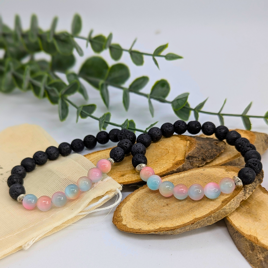 The Silver Sunbird Mermaid-Inspired Lava Stone Bracelet features black, pink, blue, and white beads displayed on rustic wooden slices, with a beige pouch and green foliage in the background.